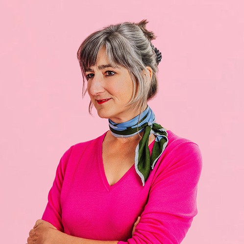 Woman with gray hair wearing a pink top and patterned scarf, standing with arms crossed against a soft pink background.