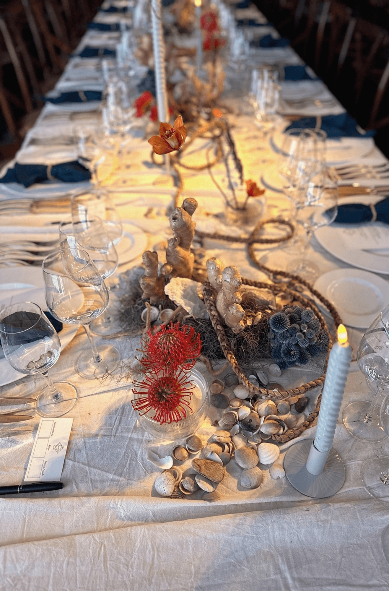 Mesa de banquete con camino de mesa de jengibre, velas blancas, elementos marinos y servilletas azul marino.