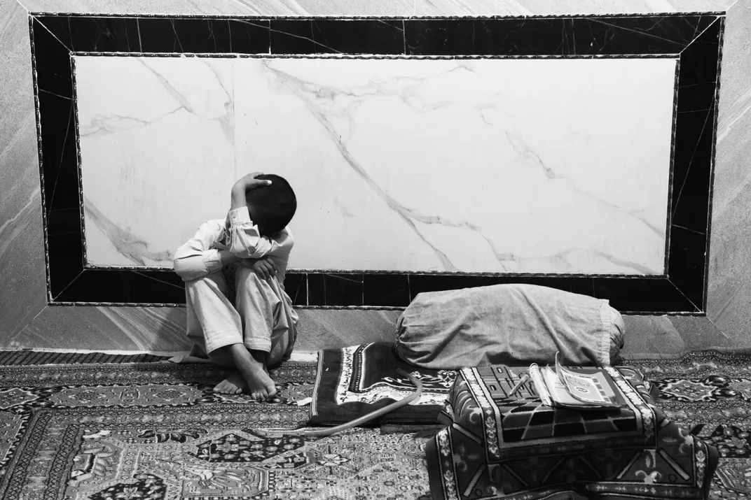 A barefoot child sitting curled with head down against a marble wall beside a sleeping figure on a patterned rug, black and white