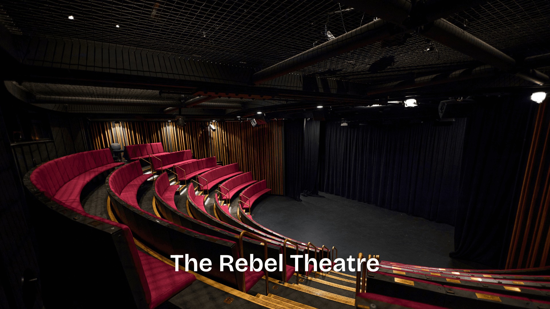 A curved, tiered theatre space with red seating and a black stage, equipped with overhead lighting and full performance curtains.