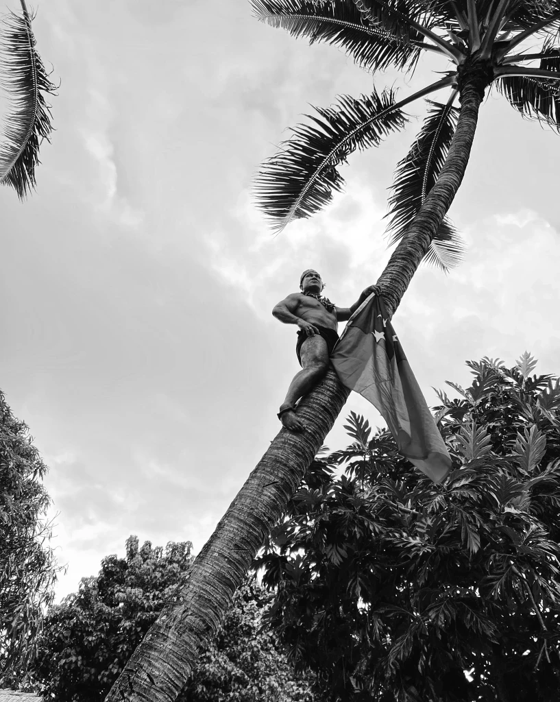 Samoan man climbing a palm tree holding the national flag of Samoa in a tropical South Pacific island landscape