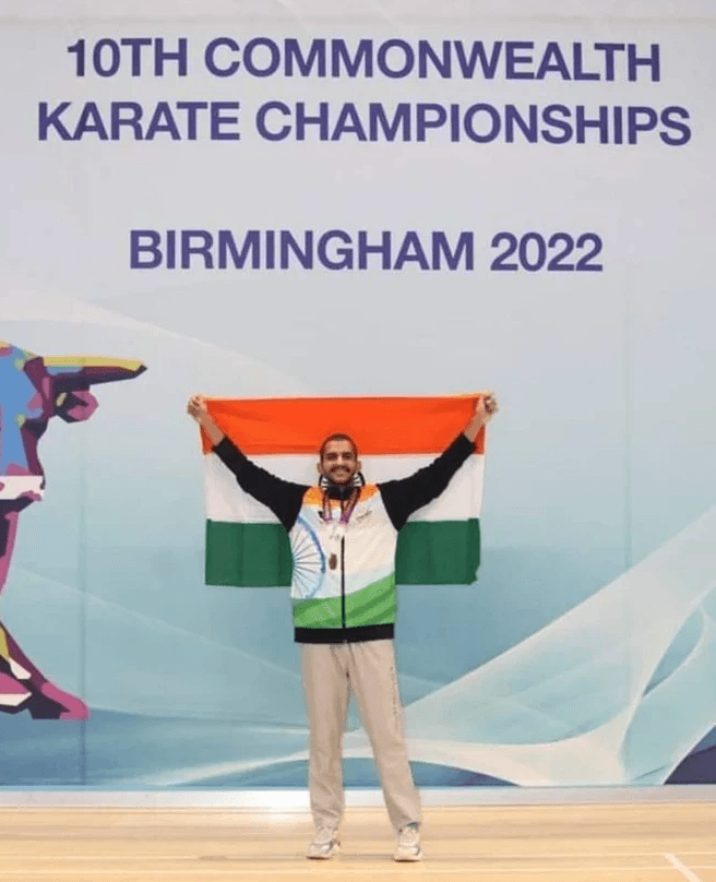 A karate athlete joyfully holds the Indian national flag overhead in front of a championship banner.