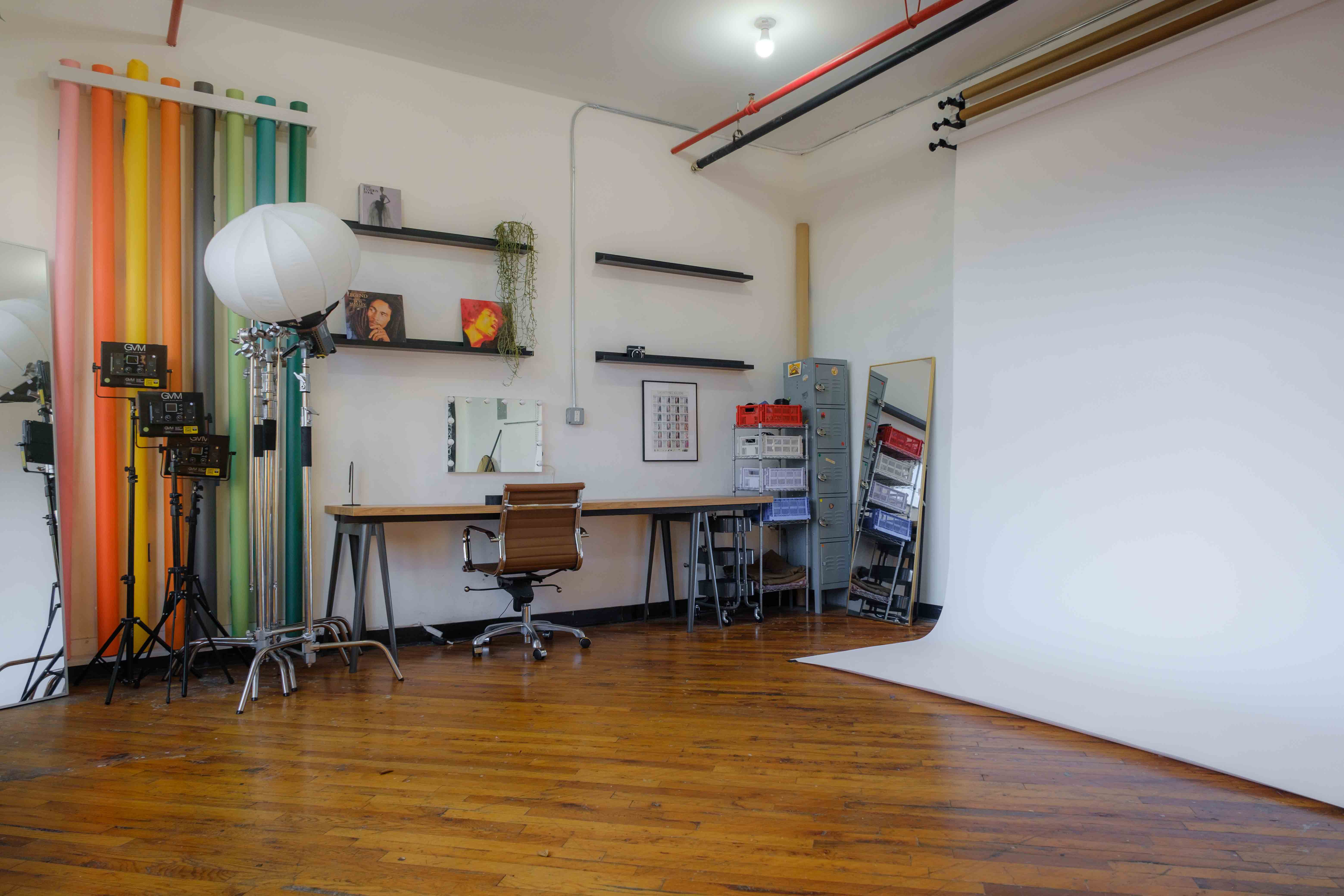 A daylight photography studio setup featuring a large white backdrop roll, a wooden desk with an ergonomic chair, and professional lighting equipment. Colorful backdrop rolls, shelves with decor, and a storage area add to the functional and creative workspace.