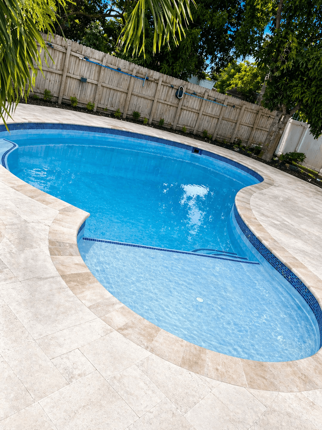 Curved residential swimming pool with beige marble coping, light stone paver deck, and wood privacy fence