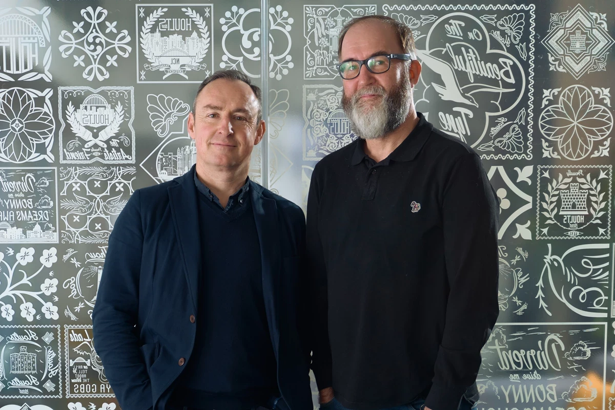 Two people stand side by side in front of a decorative glass wall featuring intricate graphic patterns. Both are wearing dark clothing in an indoor setting.