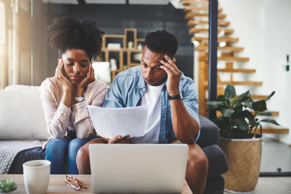 A couple sits on a couch reviewing financial documents with concerned expressions, laptop open in front of them. The image represents the stress of navigating home loans and the need for trusted guidance from Chris Lewis Home Loans.