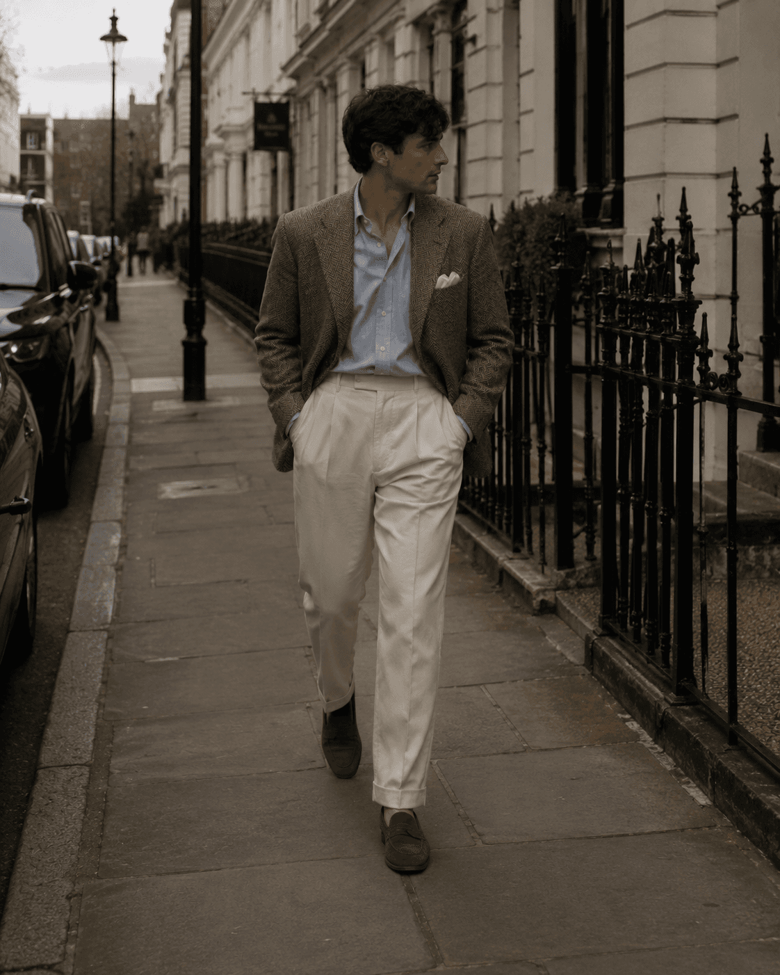 Full-length view of a man walking down a city sidewalk in classic men's smart-casual tailoring, wearing a brown textured tweed blazer with a white pocket square, a light blue button-down shirt, high-waisted off-white pleated trousers, and brown suede loafers, with elegant townhouses in the background.
