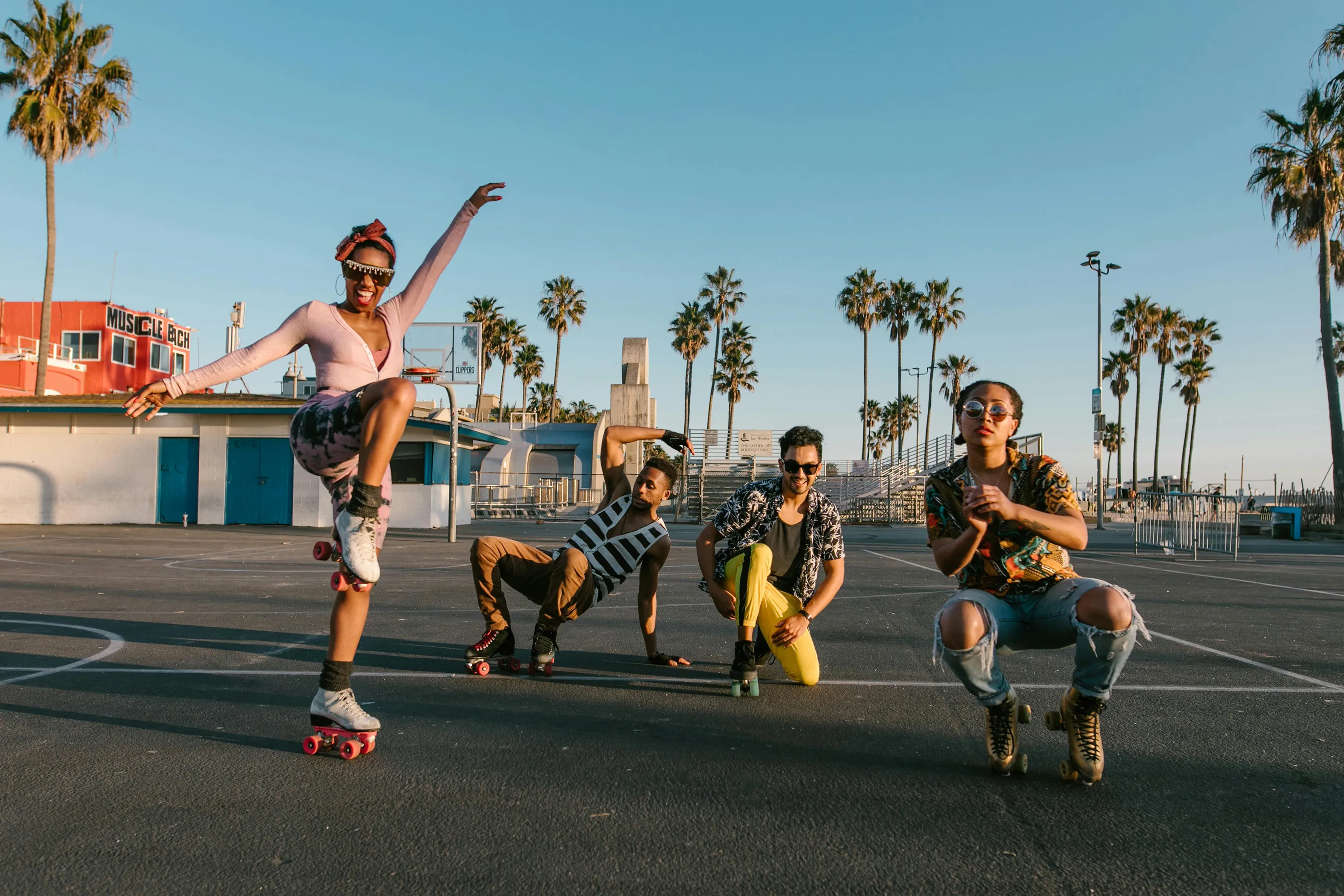 Four friends on roller skates strike dynamic poses on an outdoor court lined with palm trees