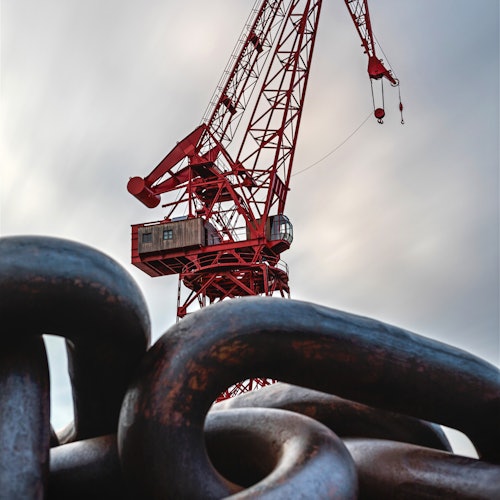 Red industrial crane with a cloudy sky background, large metal chain links in the foreground.