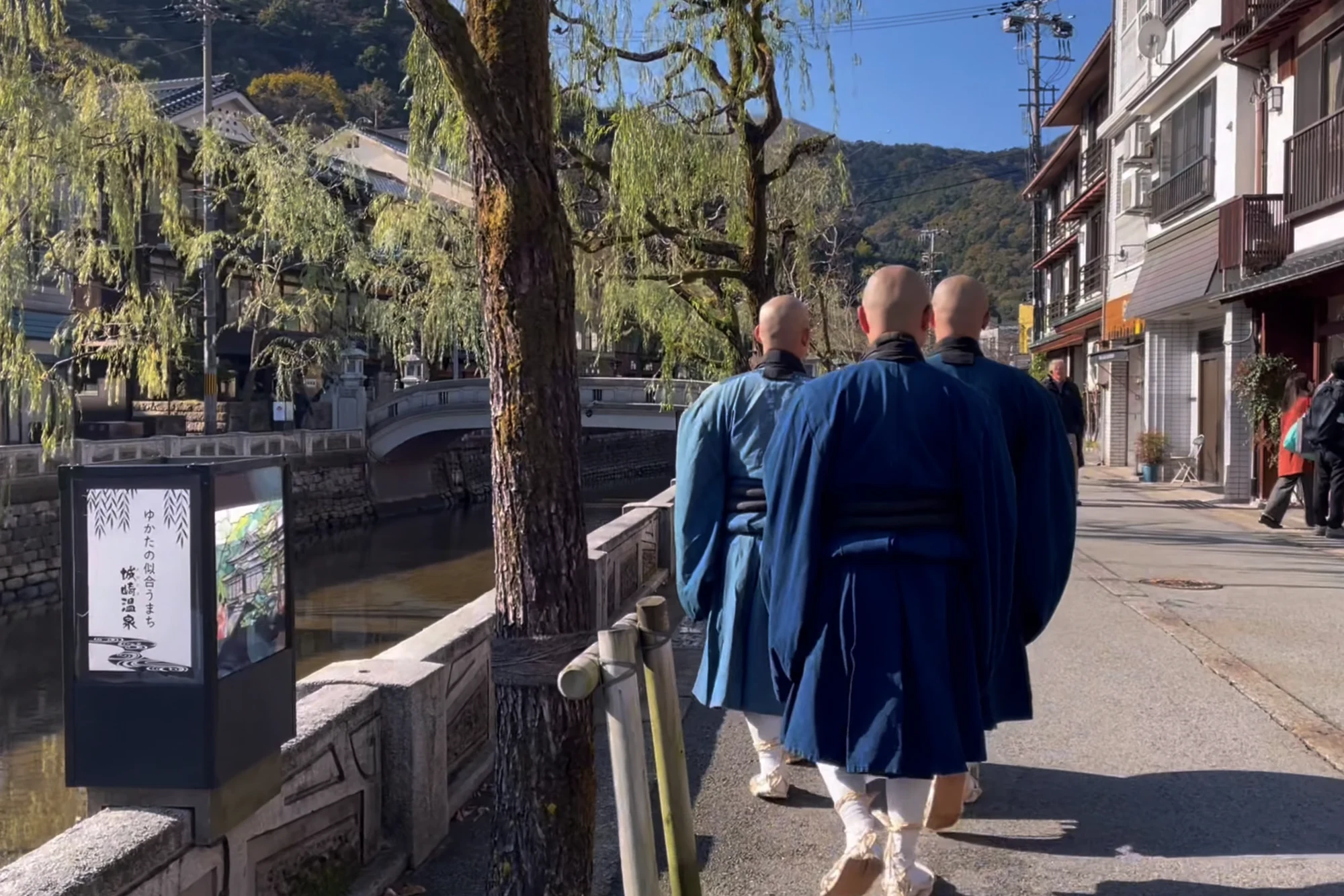 Three priests in blue robes walking together along the Kinosaki canal past an information board.