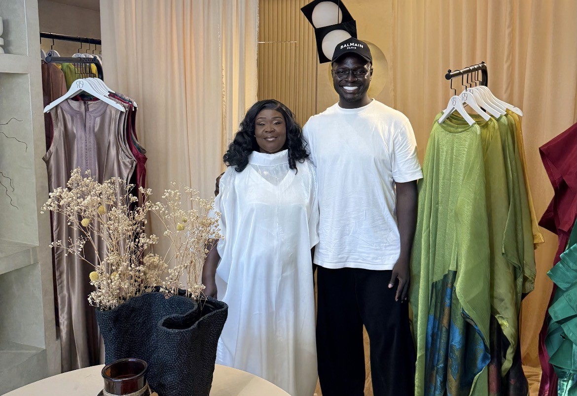 Two men sit at a wooden table with laptops, under a large red lamp. A woman walks by a window. The scene is calm, set against a red-brown wall.