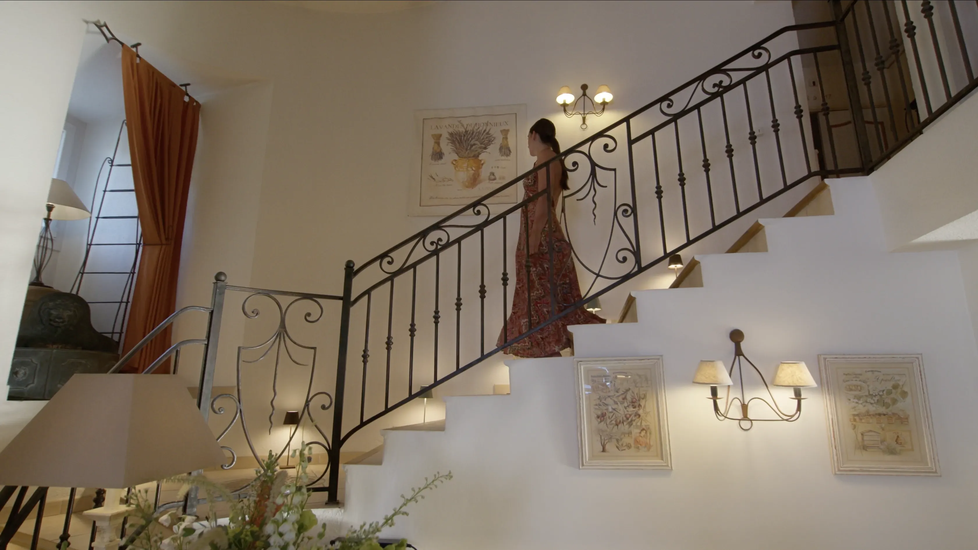 A woman in a long dress gracefully ascends a stylish staircase with ornate wrought iron railings in a well-lit, elegantly decorated home shown.