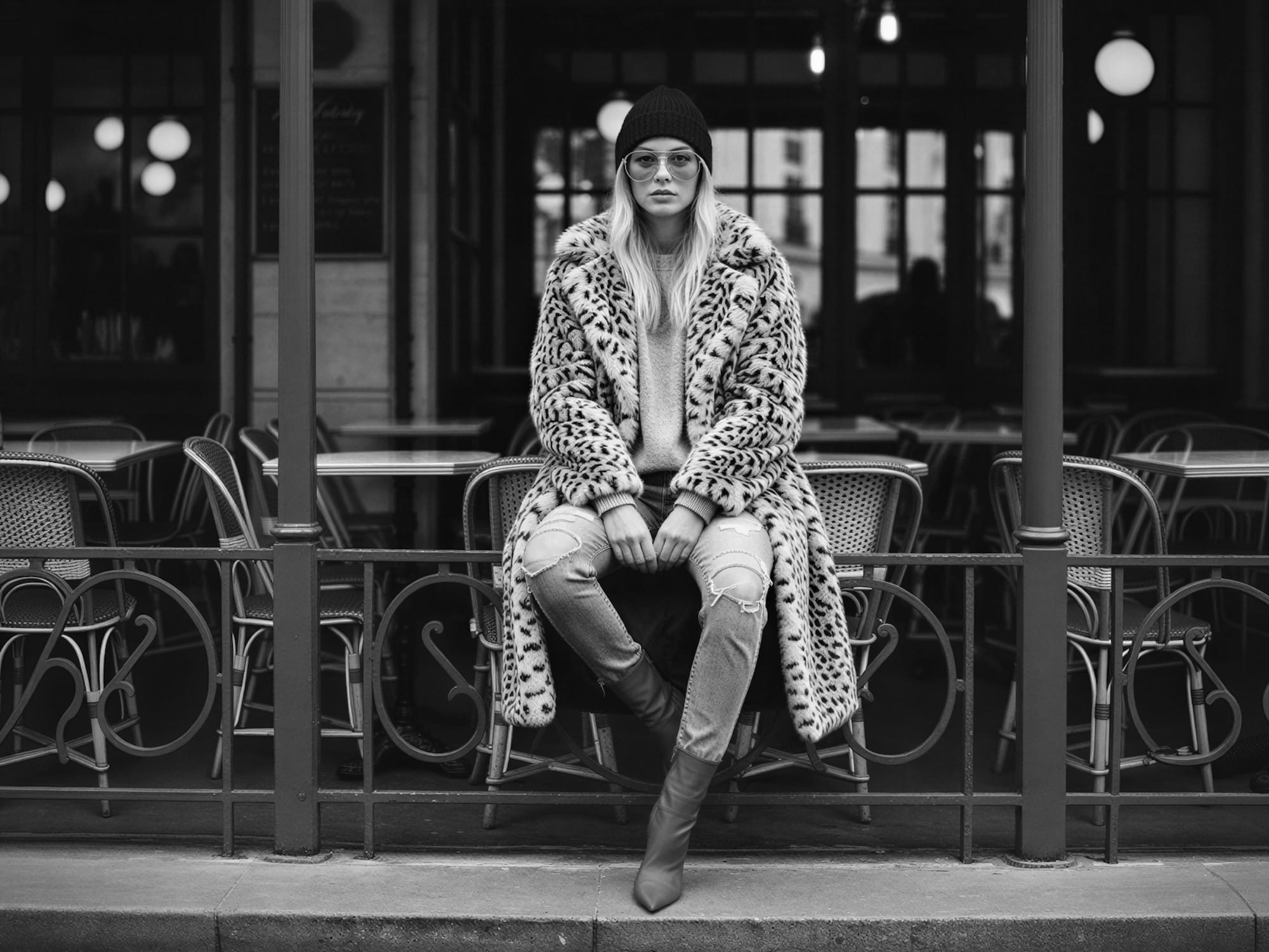 Black-and-white street-style portrait of a woman wearing a beanie, patterned coat, jeans, and boots, seated outside a café.