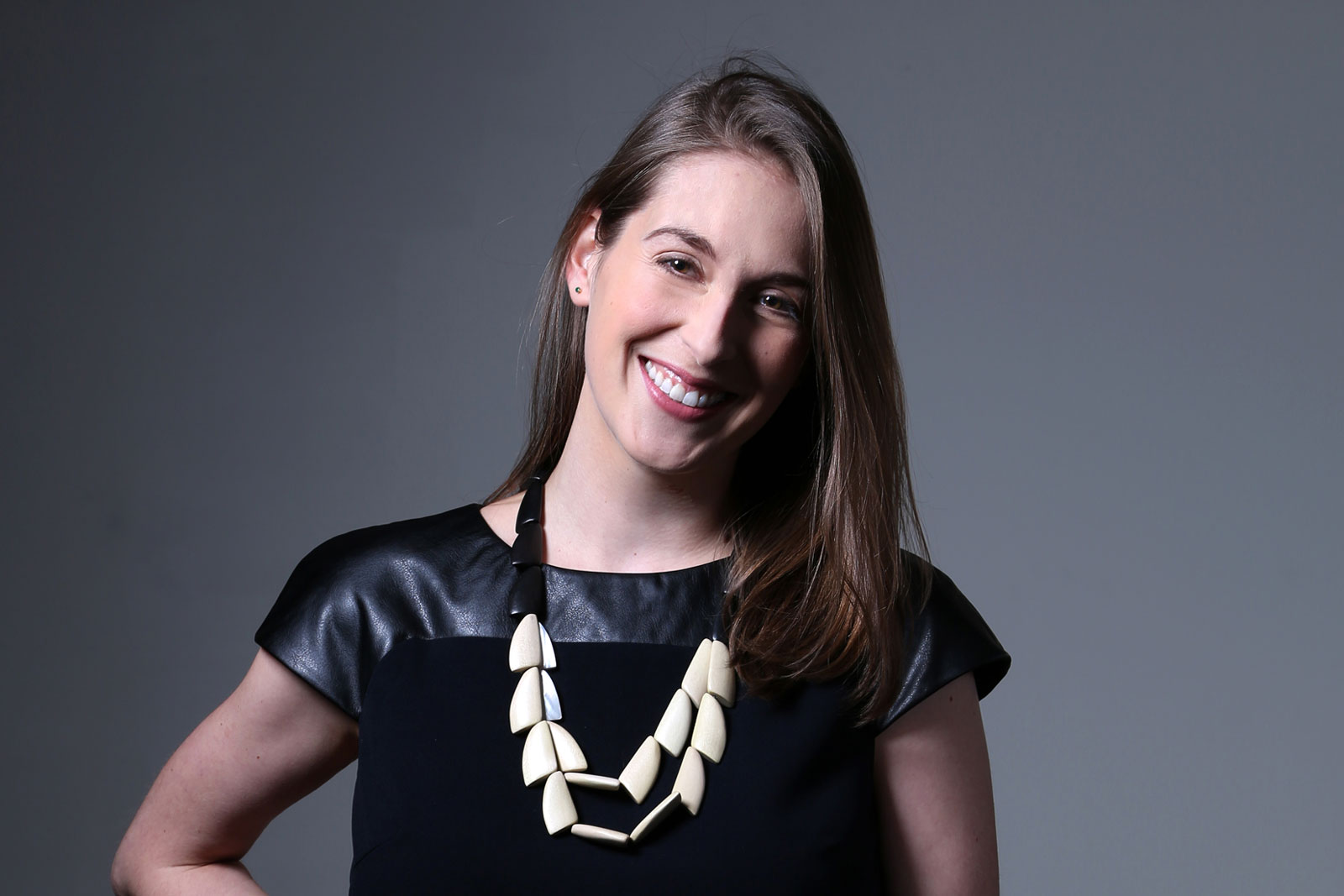 A woman with long brown hair is smiling at the camera. She is wearing a black top with shiny, leather-like sleeves and a black necklace made of off-white, geometric shapes. She is positioned against a solid gray background.
