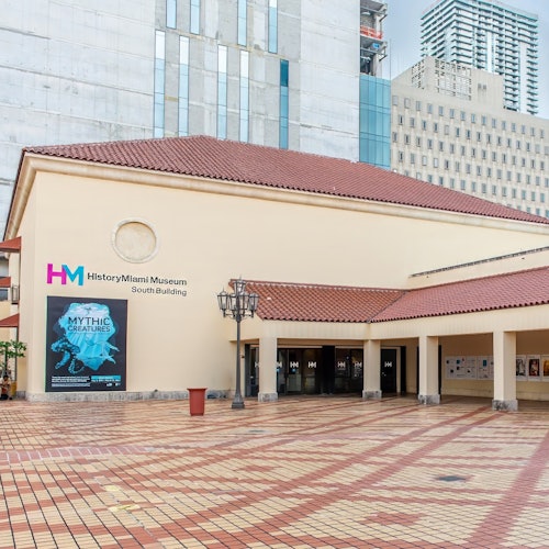 迈阿密历史博物馆南楼的入口，设有红色瓦屋顶，旁边是“神话生物”展览的海报，以及毗邻的高层建筑。