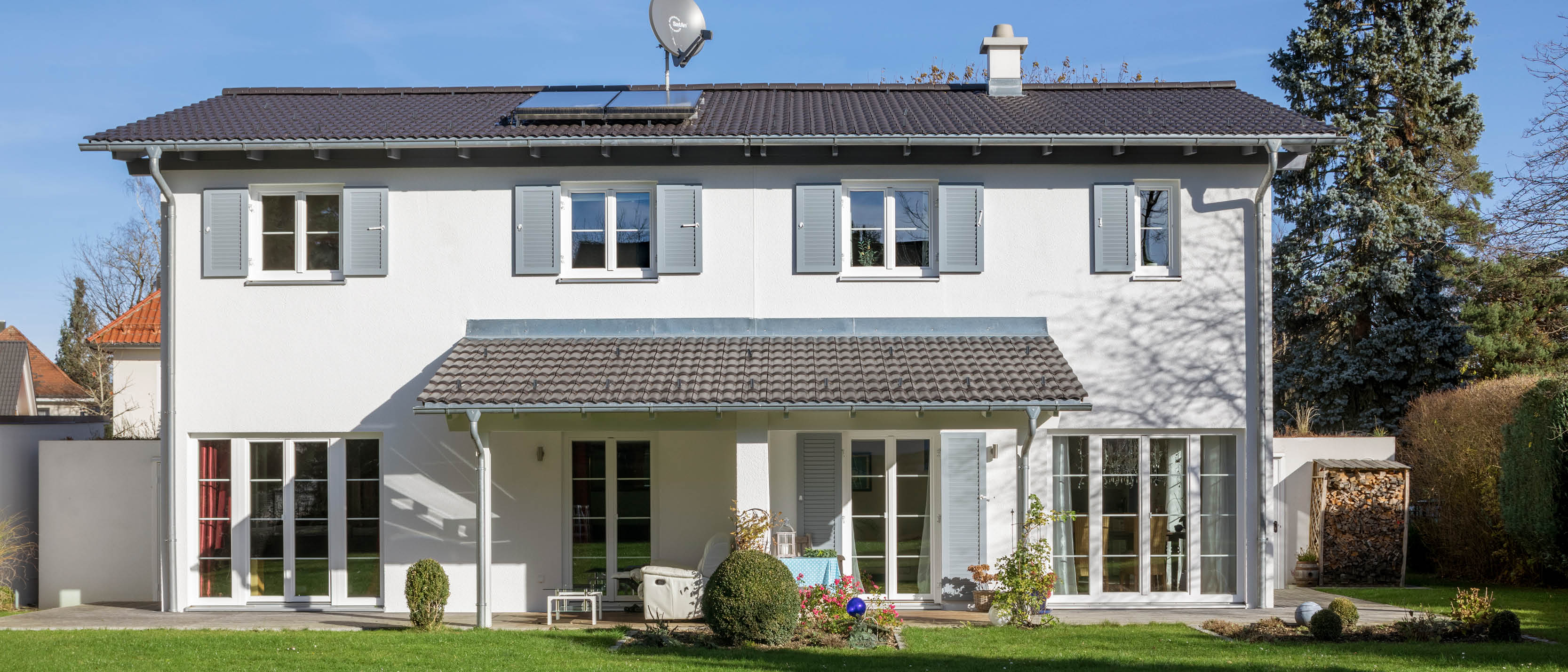 Zweifamilienhaus mit weißen Wänden, grauen Fensterläden und anthrazitfarbenem Dach, Satellitenschüssel auf dem Dach, davor grüner Rasen.