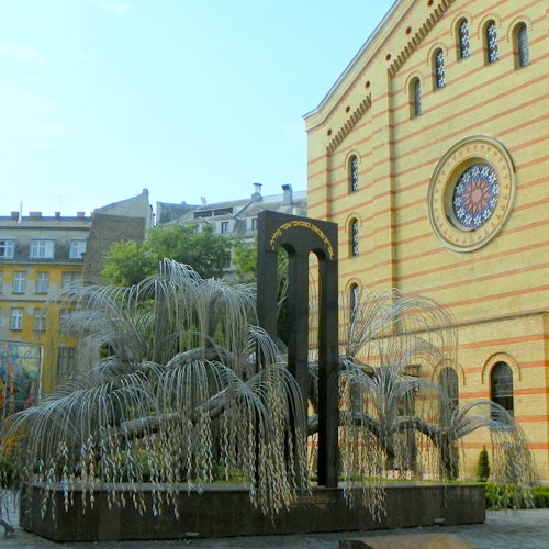 Una scultura di un salice piangente con foglie di metallo si trova accanto a un grande edificio a strisce con una finestra circolare in vetro colorato.