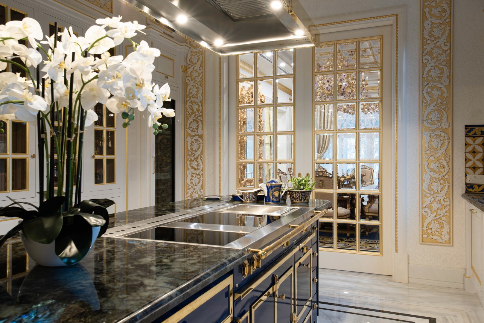 Luxurious kitchen featuring a sleek black marble countertop island with elegant gold accents, surrounded by ornate golden wall patterns, and a display of white orchids in a vase.
