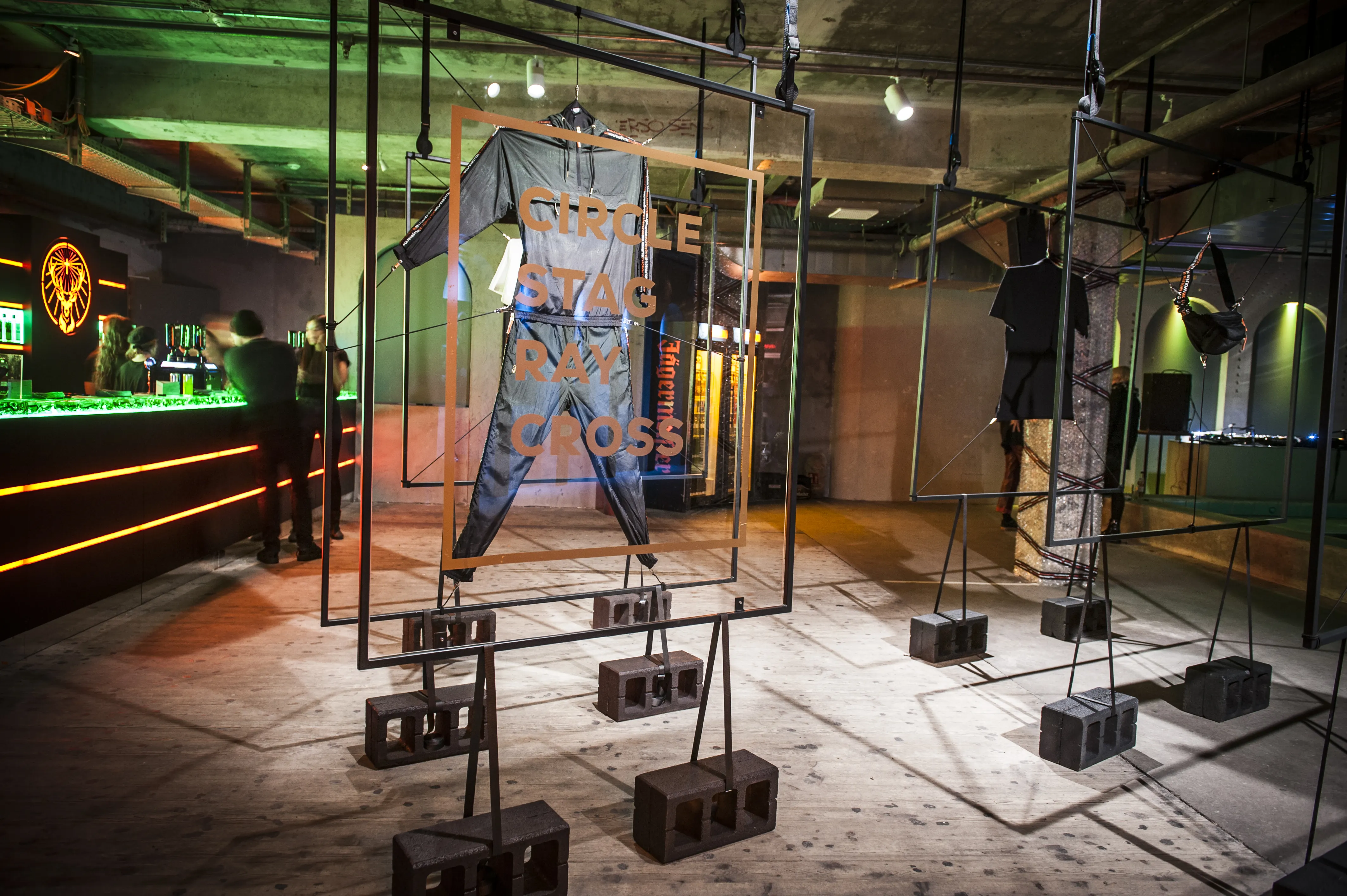 Streetwear collection at the JÄGERMEISTER launch displayed on black steel frames anchored in breeze blocks, with orange Circle Stag Ray Cross lettering and a green-lit bar.