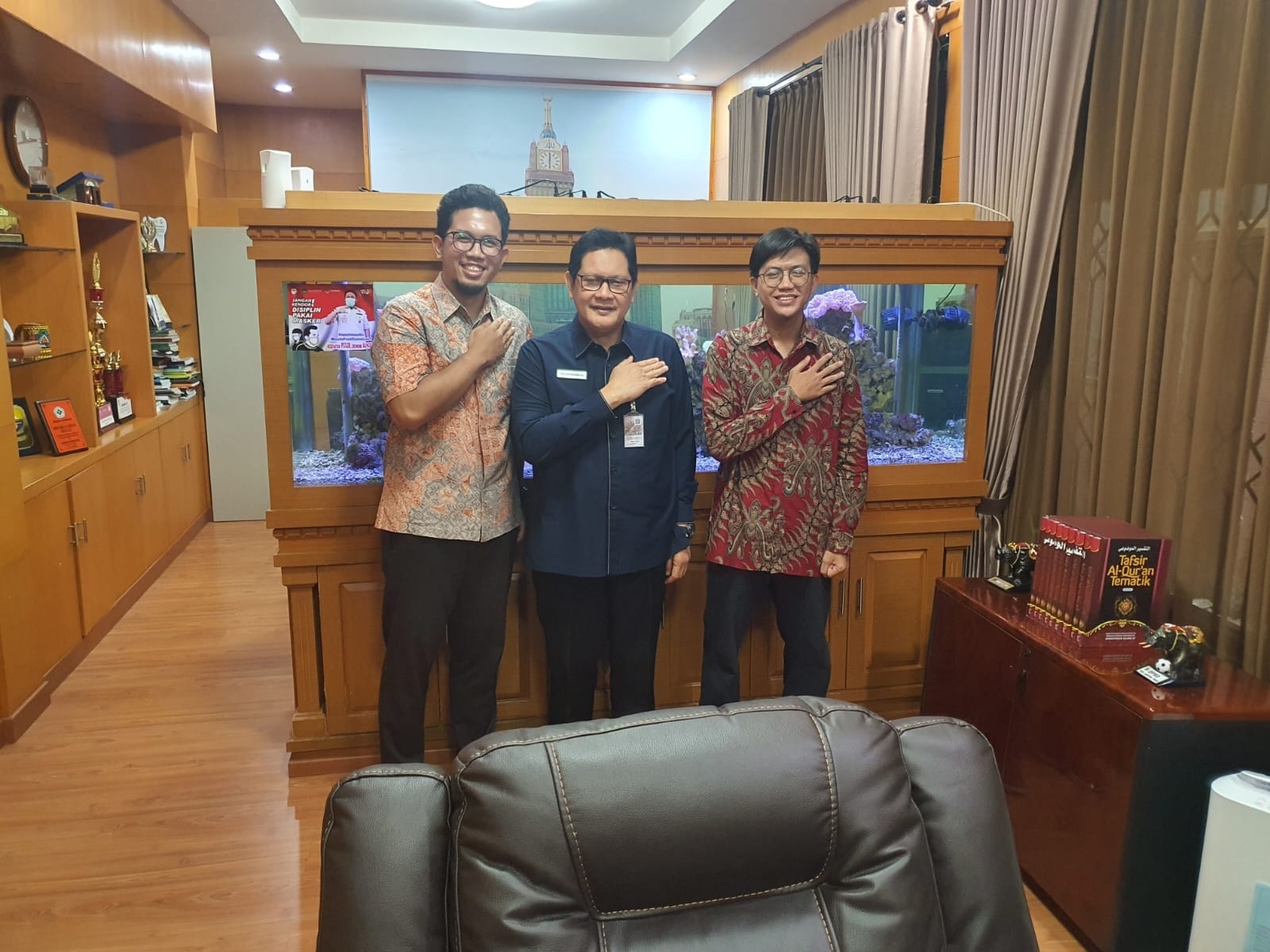 Three people posing in an office in front of a large aquarium and wood-paneled walls at the South Sumatra Health Office.