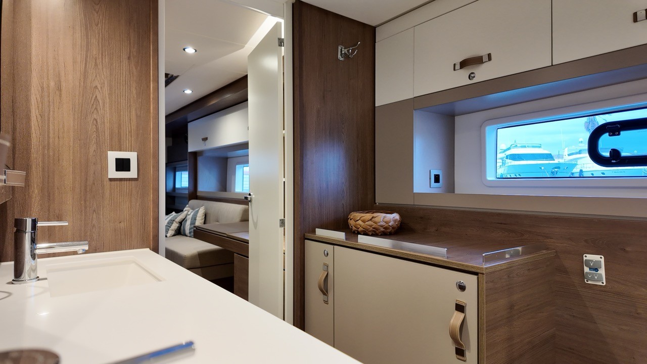 Bali 4.4 catamaran cabin and head layout view showing the en-suite basin with chrome tap, open doorway leading to a double cabin with upholstered berth and striped cushions, walnut cabinetry throughout, hull portlight window with marina view, and smart home control panel on the bulkhead