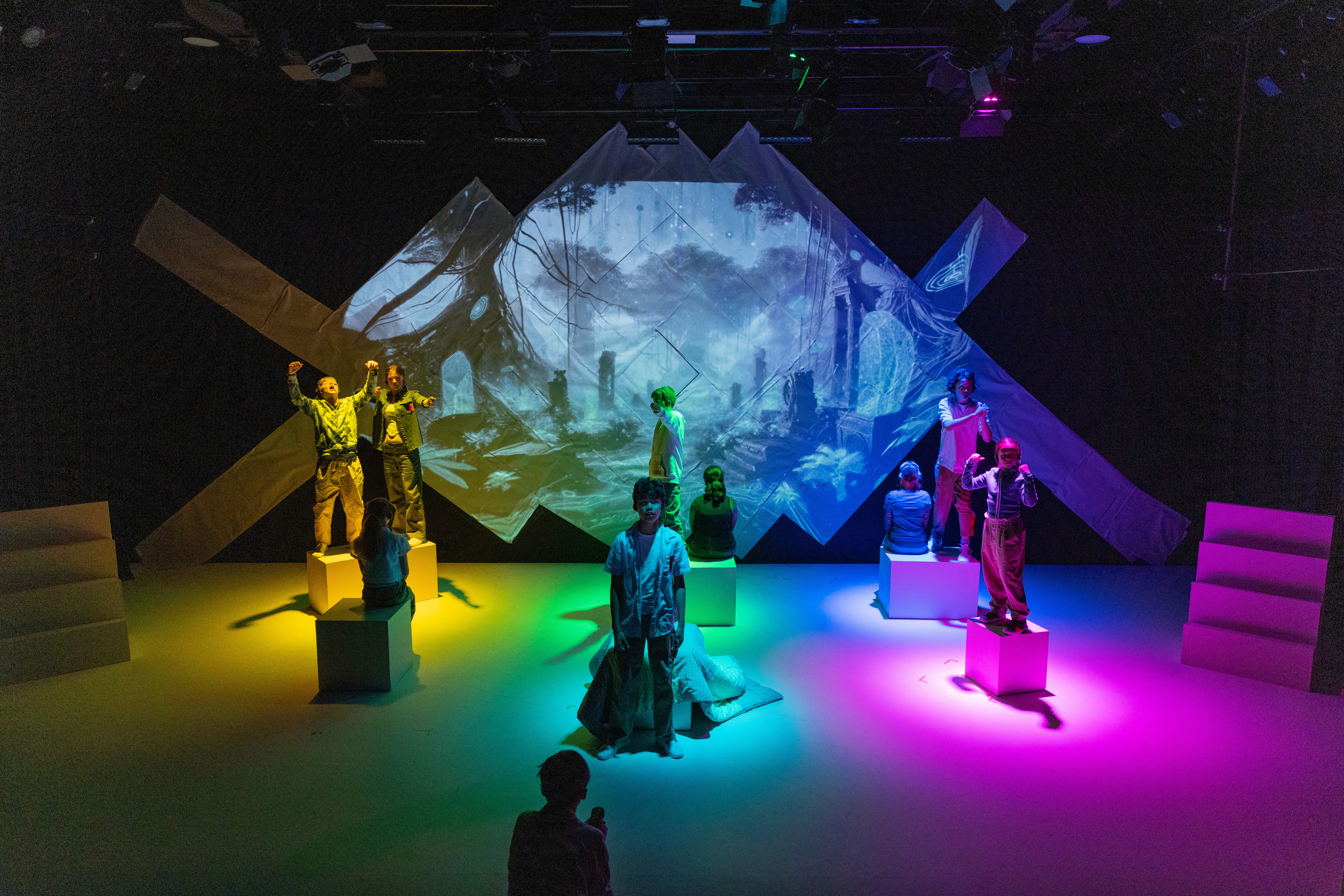 Performers stand on white boxes across a wide stage as vivid multicoloured lighting and an immersive fantasy landscape projection fill the backdrop, creating a surreal digital world.