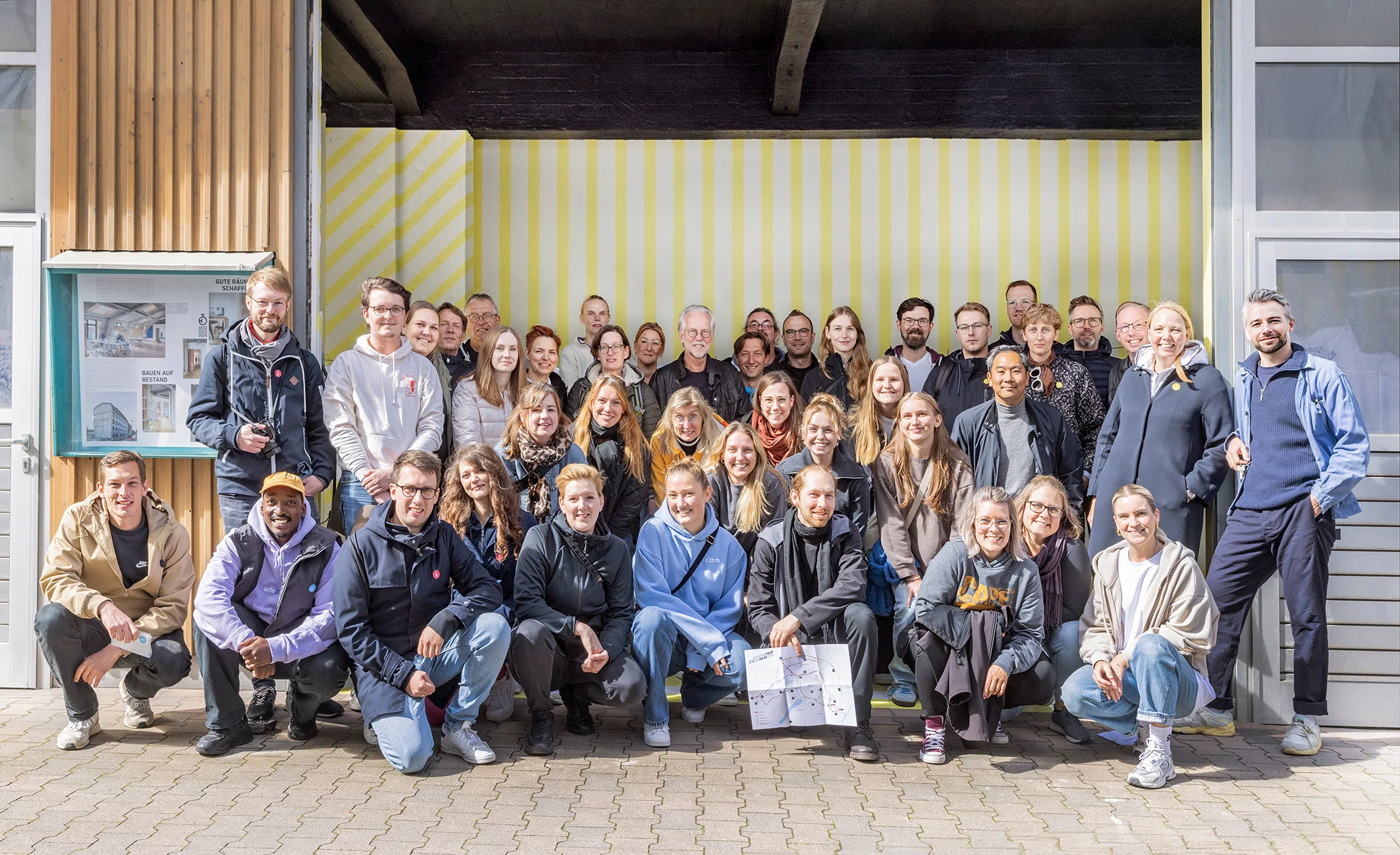 Großes Gruppenfoto des MAAS & PARTNER Teams vor einer gelb-weiß gestreiften Außenfassade, aufgenommen im Hofbereich des Bürostandorts.