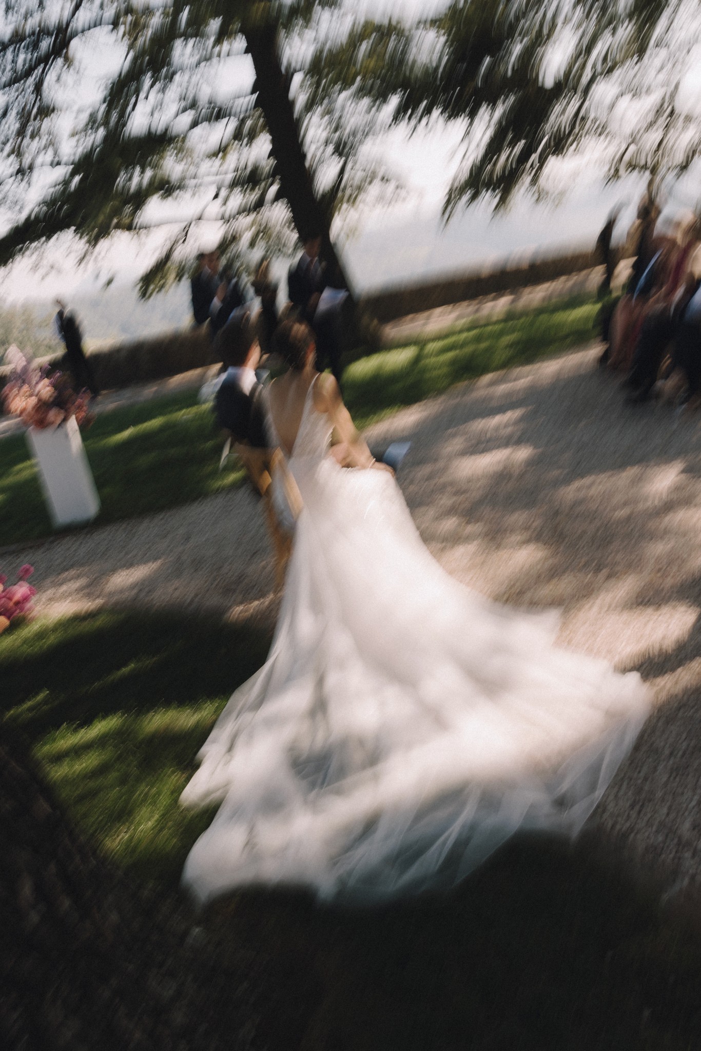 Sposa in abito bianco cammina sotto gli alberi al Castello di Tabiano, strascico fluente, luce filtrata tra le fronde
