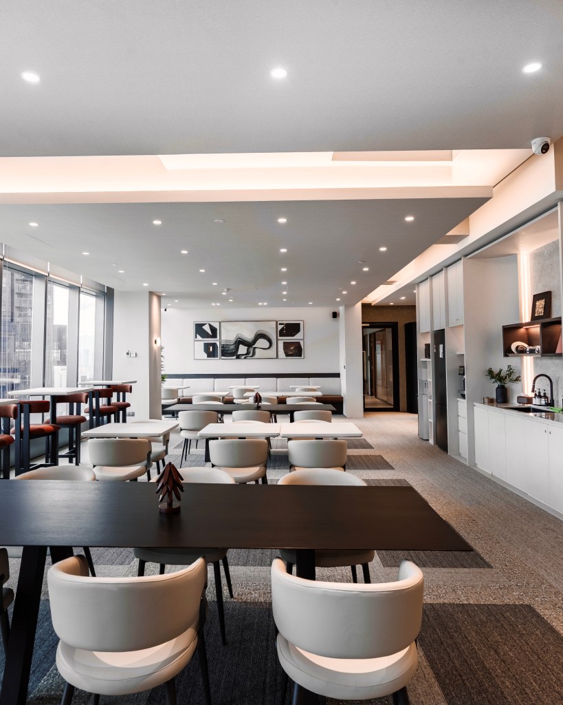 A modern coworking space featuring several white chairs around black tables, with a sleek design and minimalistic decor. Large windows provide natural light, and artworks are displayed on the walls.
