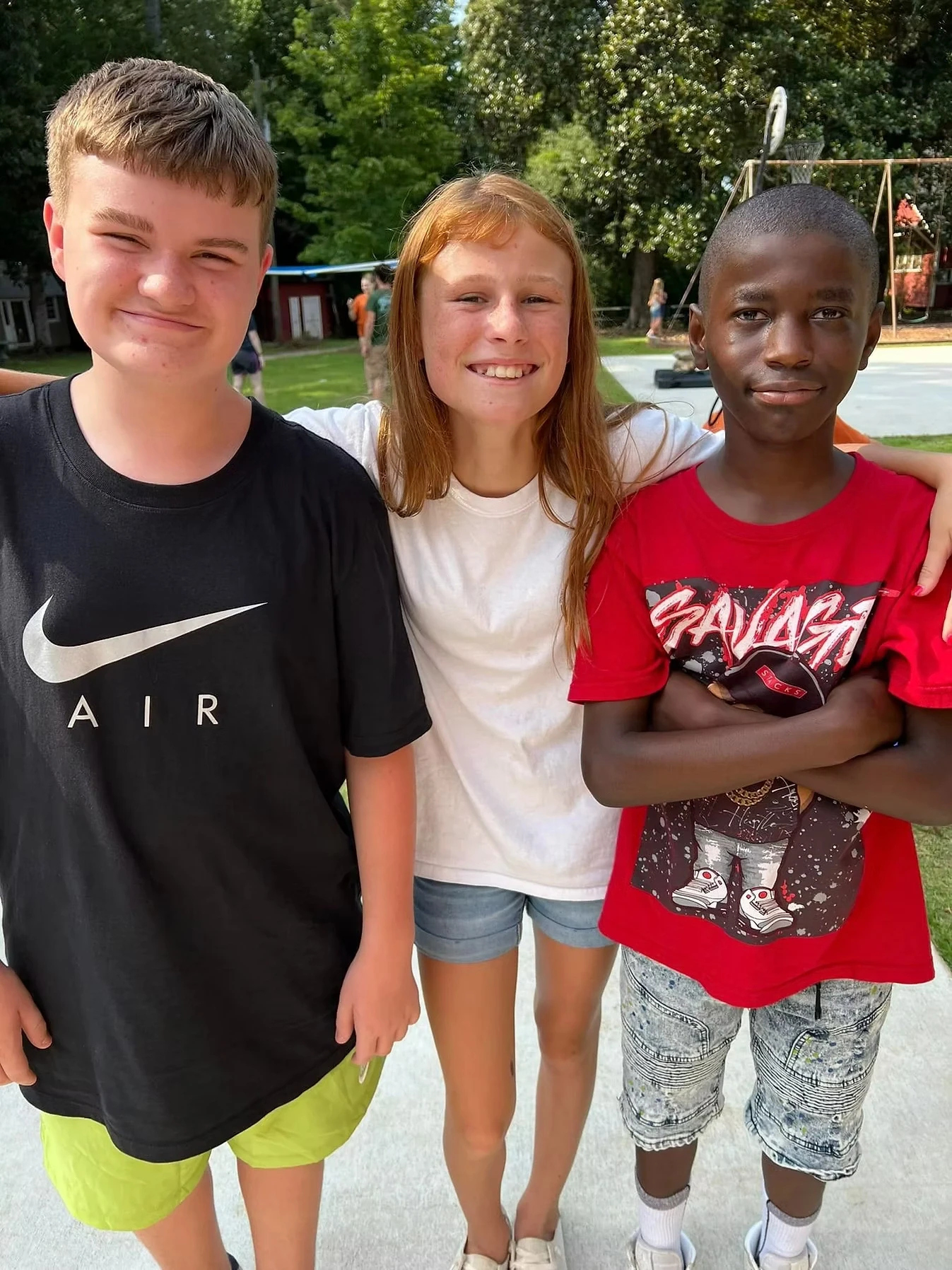 Three smiling campers, a boy and two girls, stand close together, arms around each other. They're outdoors on a paved area.