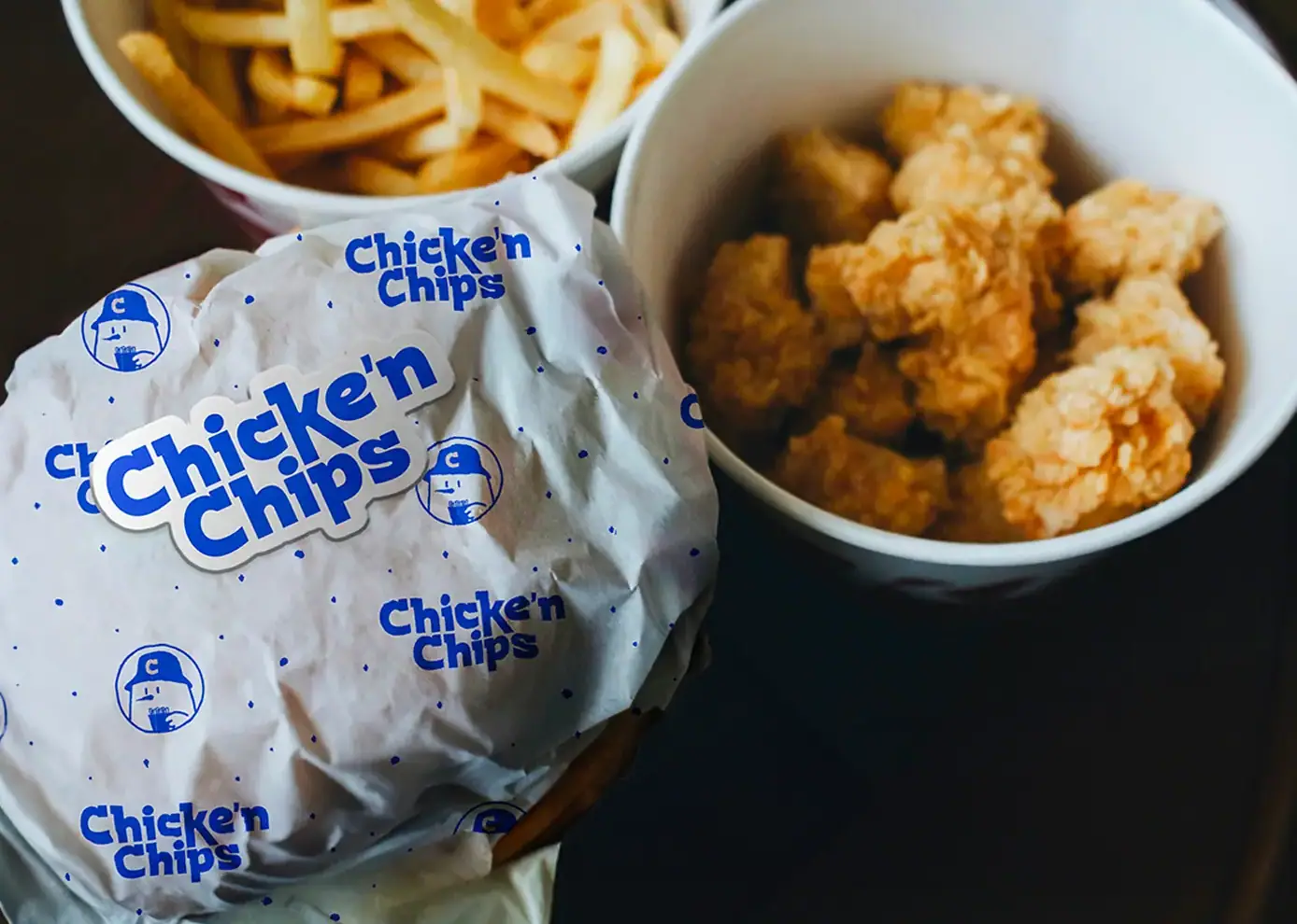 Burger emballé dans du papier d'emballage imprimé au logo Chicke'n Chips, accompagné de deux bols de frites et de nuggets de poulet frit.
