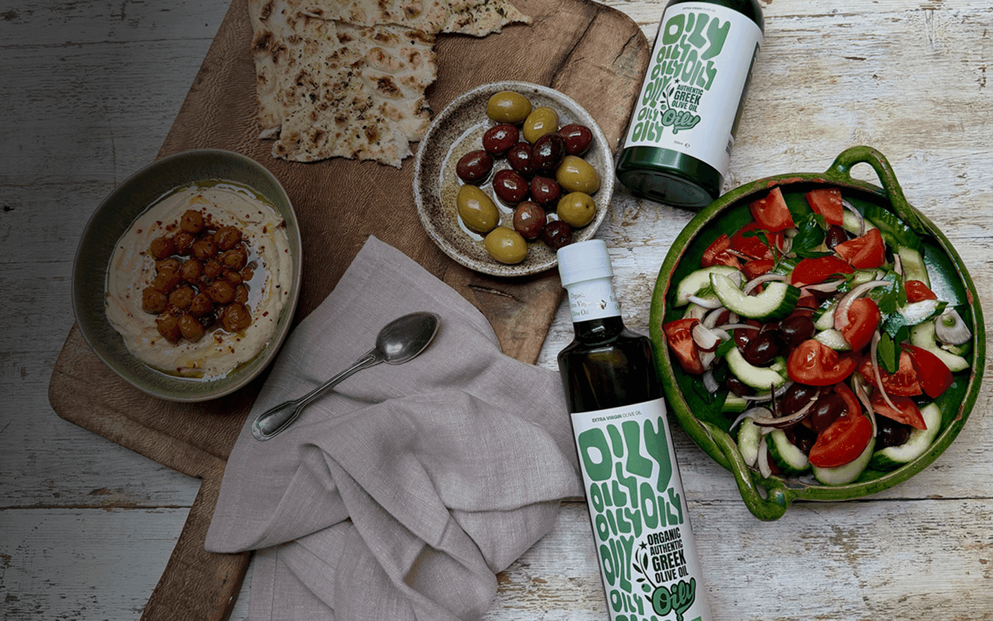 Two bottles of Oily next to a spread of hummus and greek salad