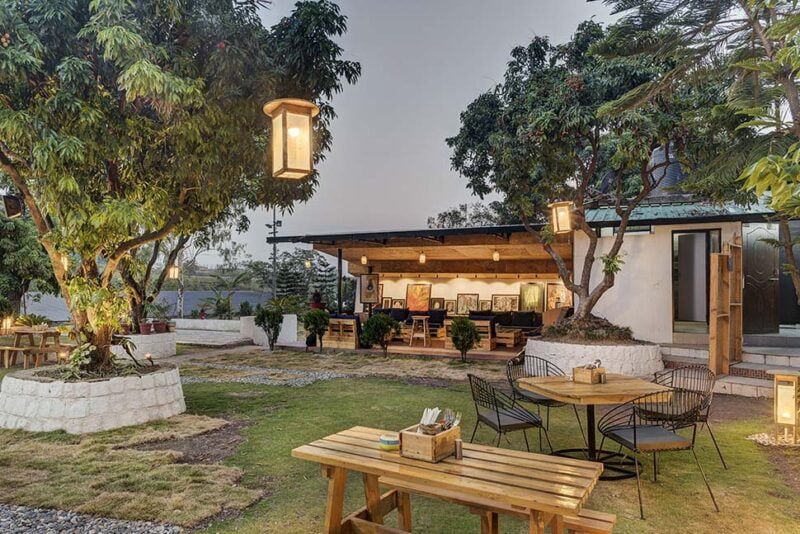 Al fresco charm: Warm lantern light illuminates this serene outdoor dining space, where rustic wooden furniture sits amidst mature trees and stone planters.