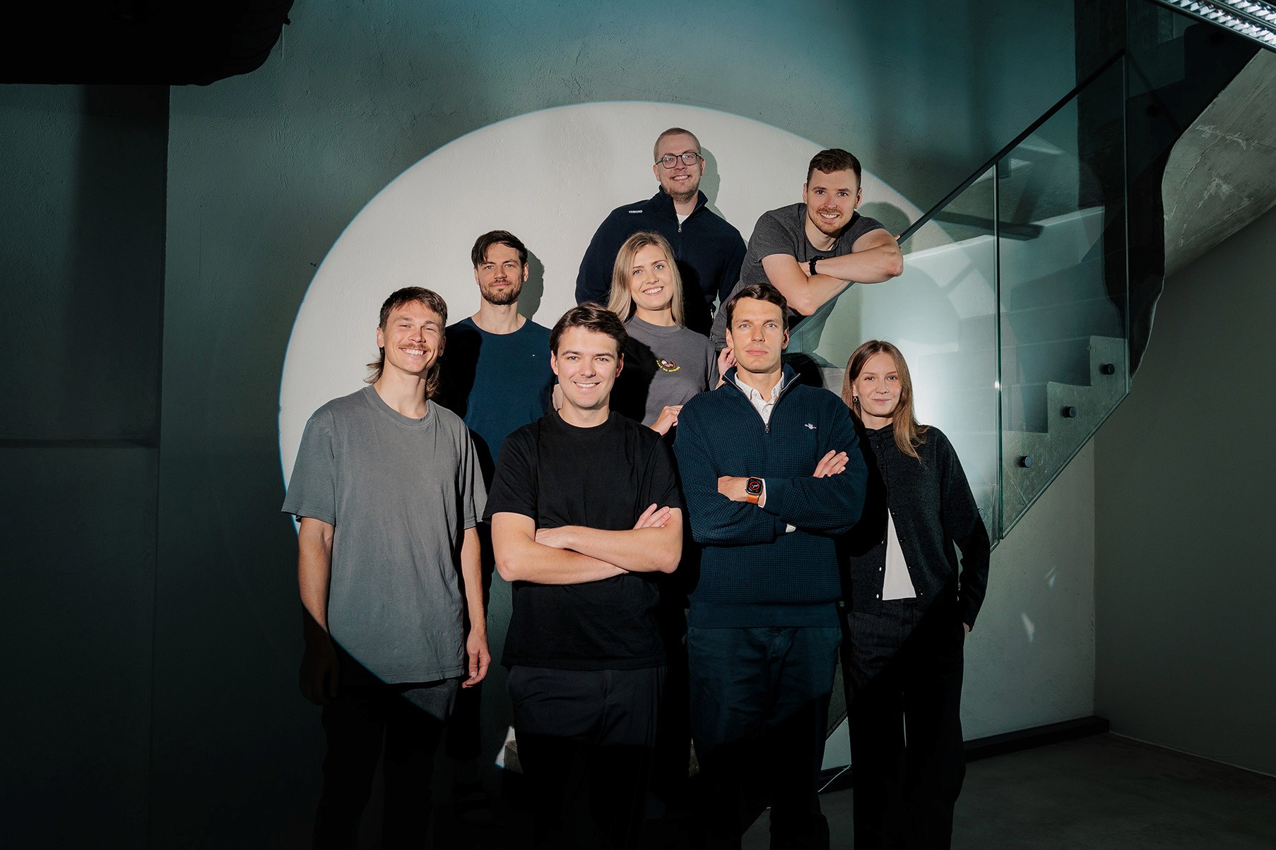 A team of eight sales outreach experts stands together inside a modern, industrial-style building, with glass railings and concrete stairs, under warm ambient lighting, conveying a sense of camaraderie and professionalism.