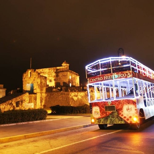 Một chiếc xe buýt du lịch hai tầng với đèn sáng được ghi nhãn "Centro Historico" đang đậu gần một pháo đài lịch sử được chiếu sáng vào ban đêm.