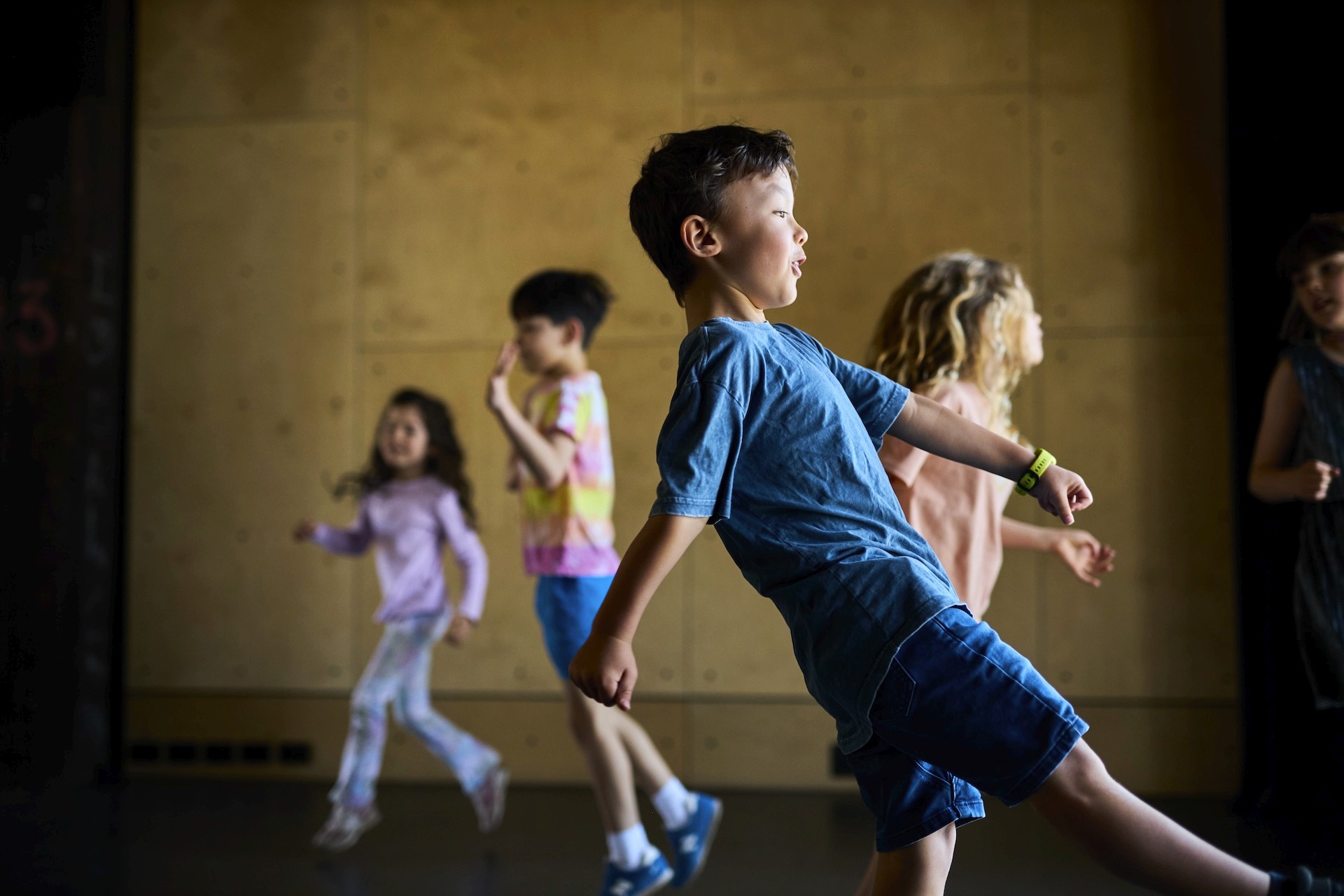 Young children run and play through a movement activity in a bright rehearsal room.