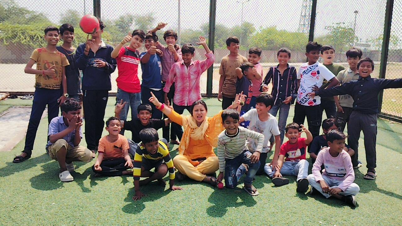 A smiling woman with her arms crossed, standing against a dark green background. She has long, dark hair.