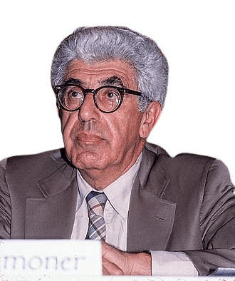 A distinguished elderly man with gray, curly hair and thick-rimmed glasses, wearing a beige suit and a striped tie, is seated at an event or conference. His expression is thoughtful as he looks slightly upward, with a neutral background suggesting an indoor, professional setting.