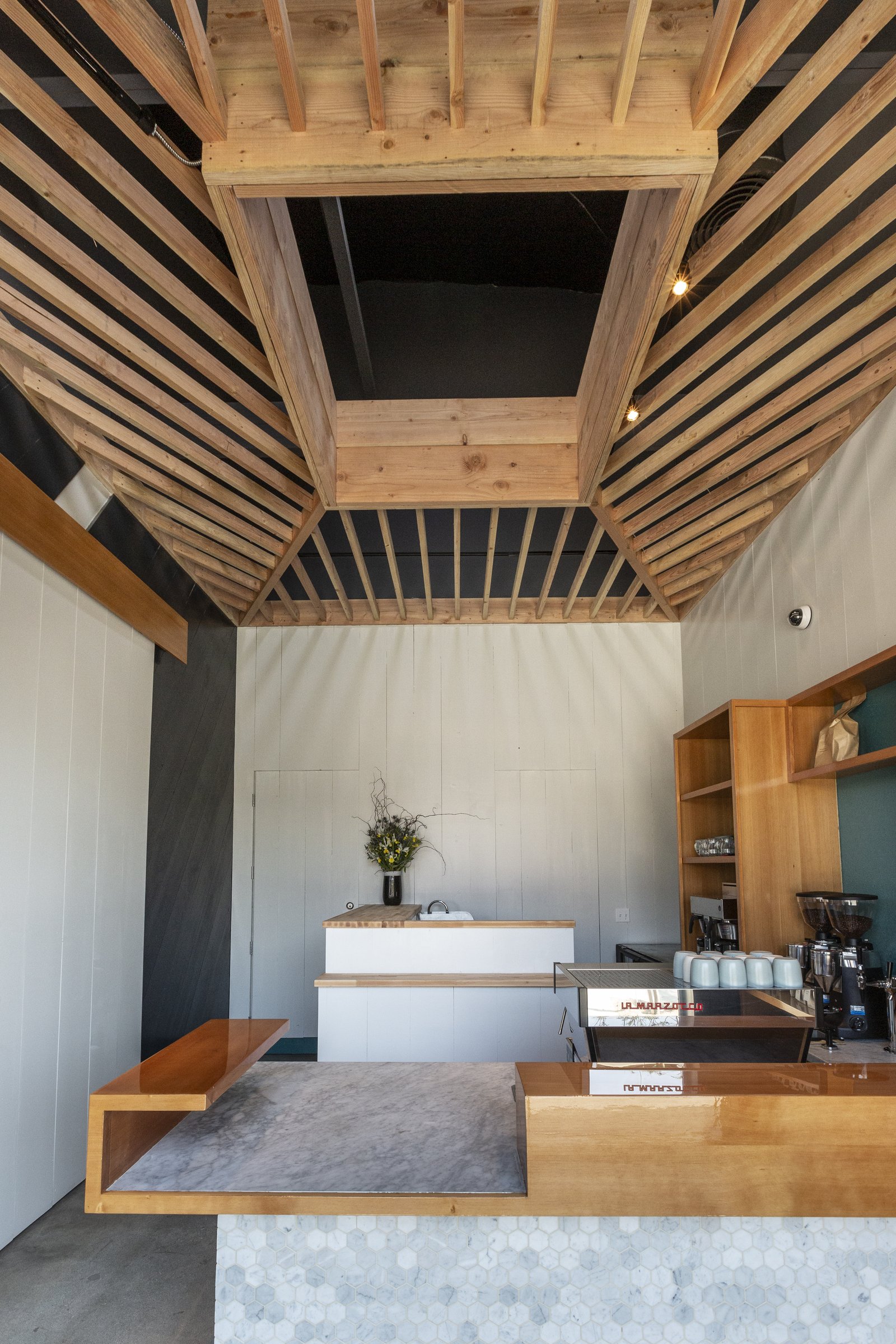 Narrow coffee bar with pale tile walls, a timber-edged counter and an open slatted timber ceiling.