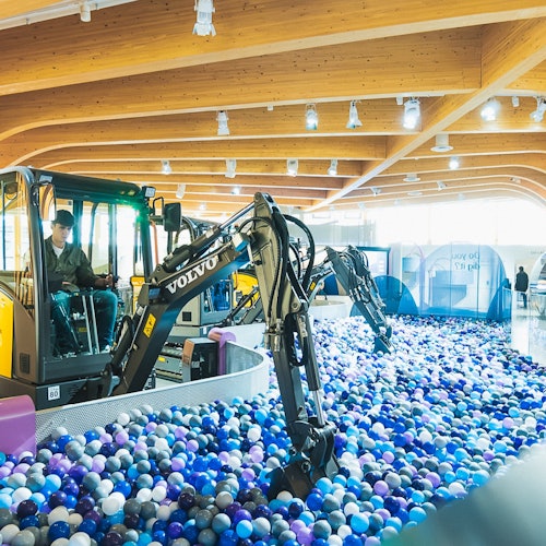 En man hanterar en minigrävare i en stor inomhus bollhavs fylld med blå, lila och vita bollar under ett trätak.