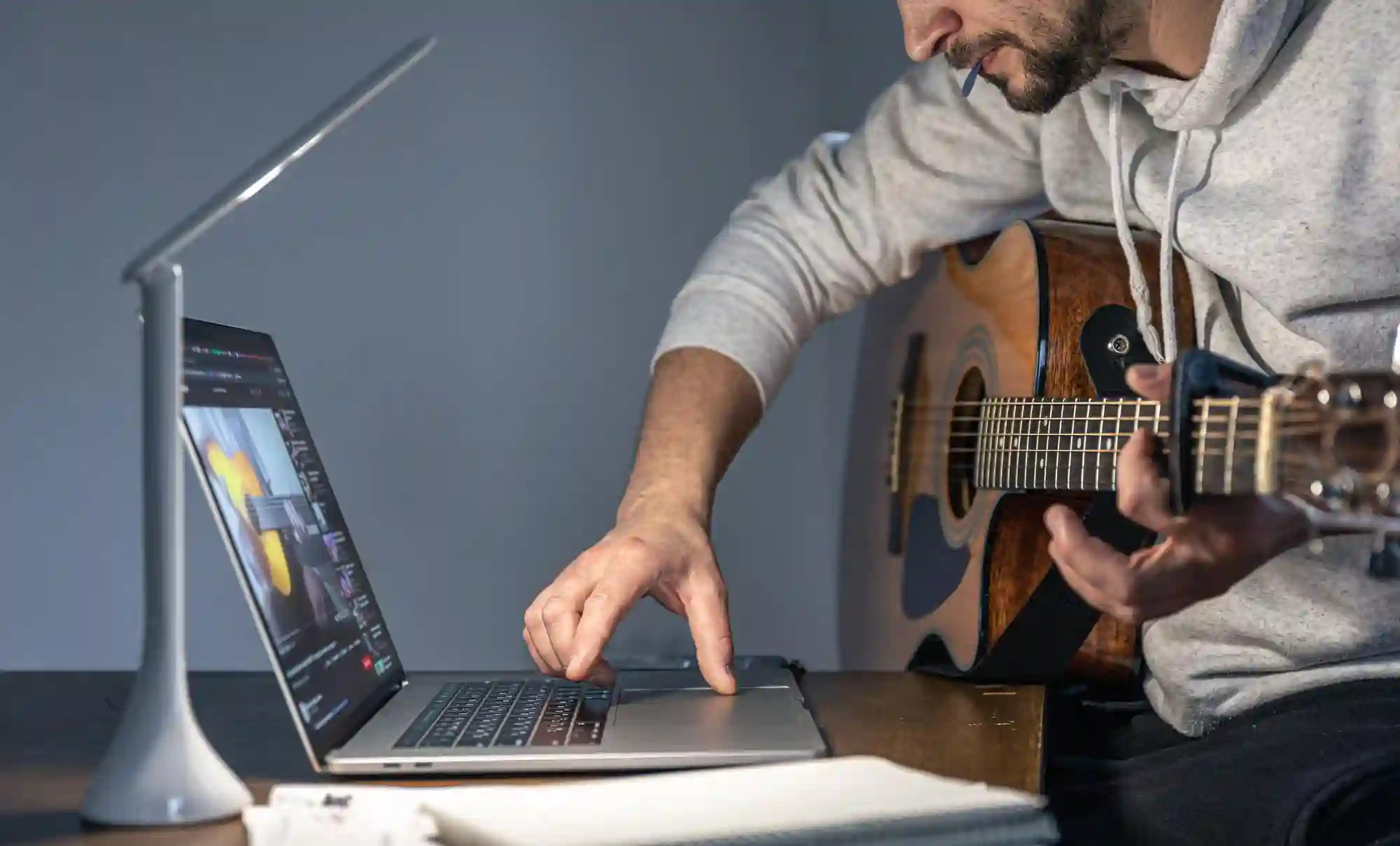 A guitarist uses a laptop and desk lamp to record music, illustrating the modern home studio setup.