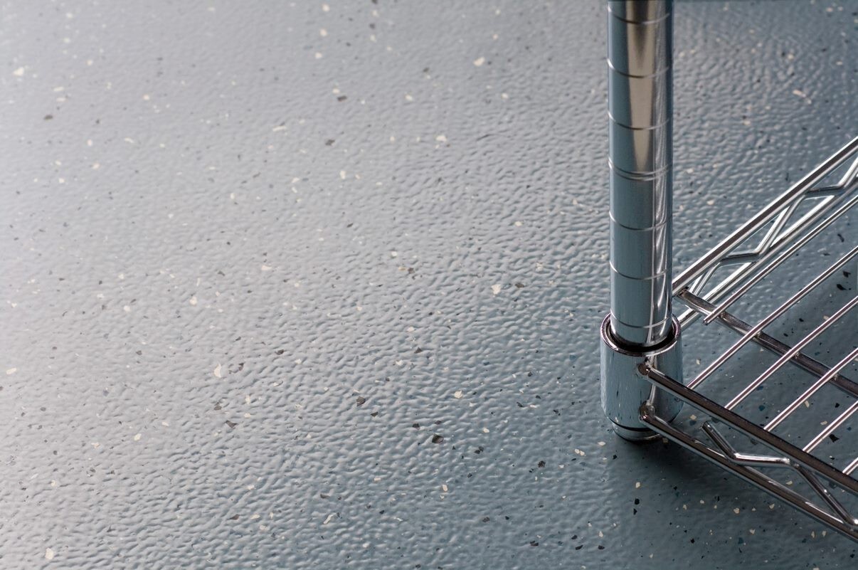 Dettaglio tecnico di una pavimentazione industriale in resina grigia con finitura antiscivolo ed effetto seminato, su cui poggia la base cromata di una scaffalatura metallica