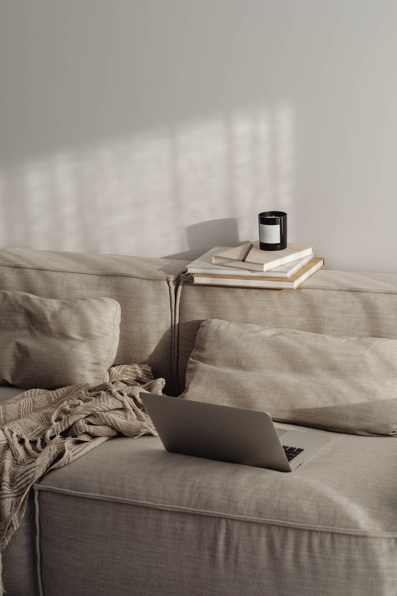 Eine graue moderne Couch steht vor einer hellen Wand, auf der Couch liegt eine gemütliche Decke und ein aufgeklappter Laptop, auf der Rückenlehne liegen mehrere Notizbücher und eine minimalistische Duftkerze