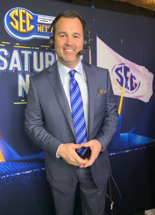 Tom Hart in a blue suit and striped tie stands smiling with a headset on in front of an SEC Network Saturday Night backdrop and waving SEC flag.