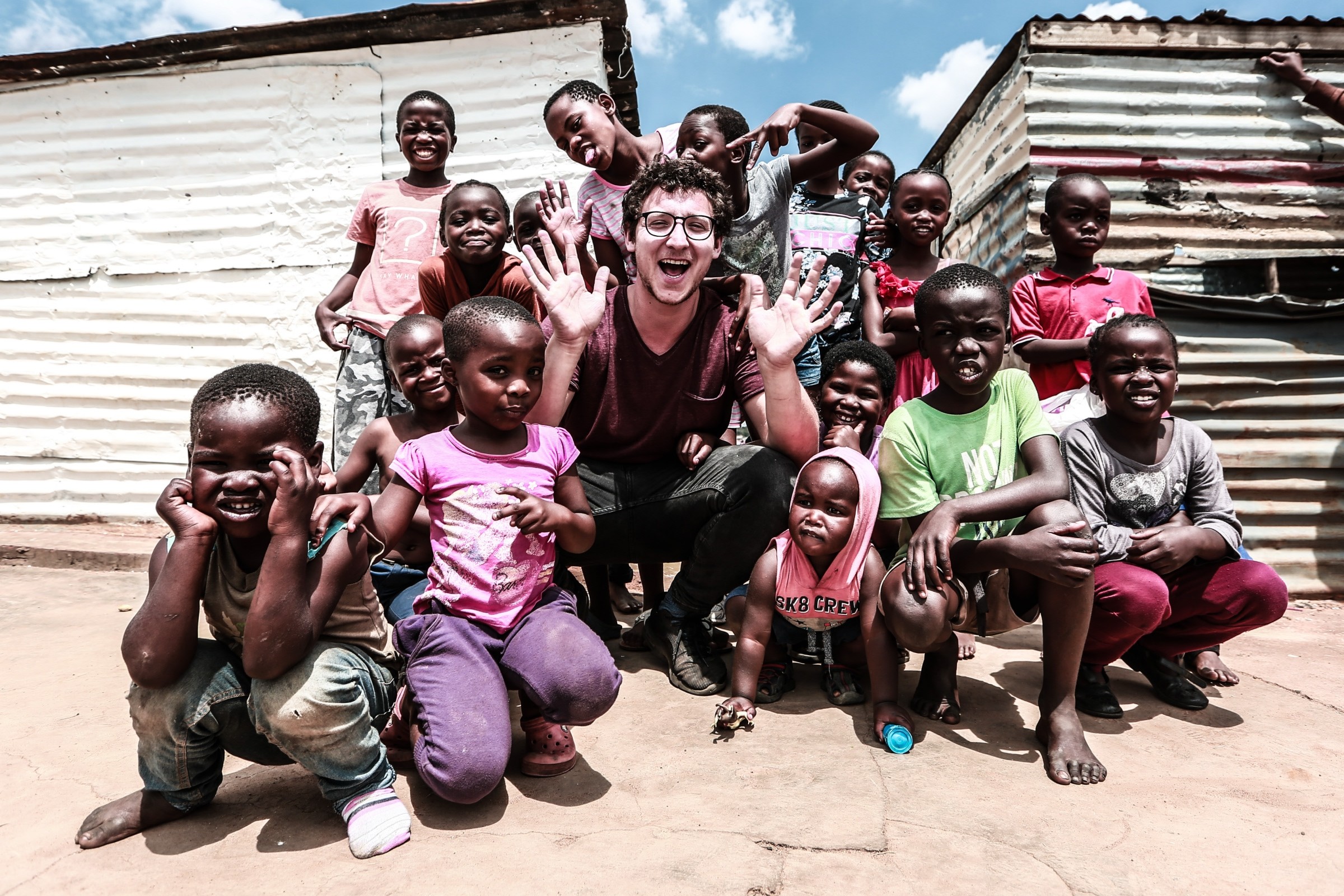 Julius Dein crouching on dusty ground in front of corrugated-metal shacks, surrounded by a group of African children of various ages, laughing with hands up some standing and some seated, all smiling and looking toward the camera.