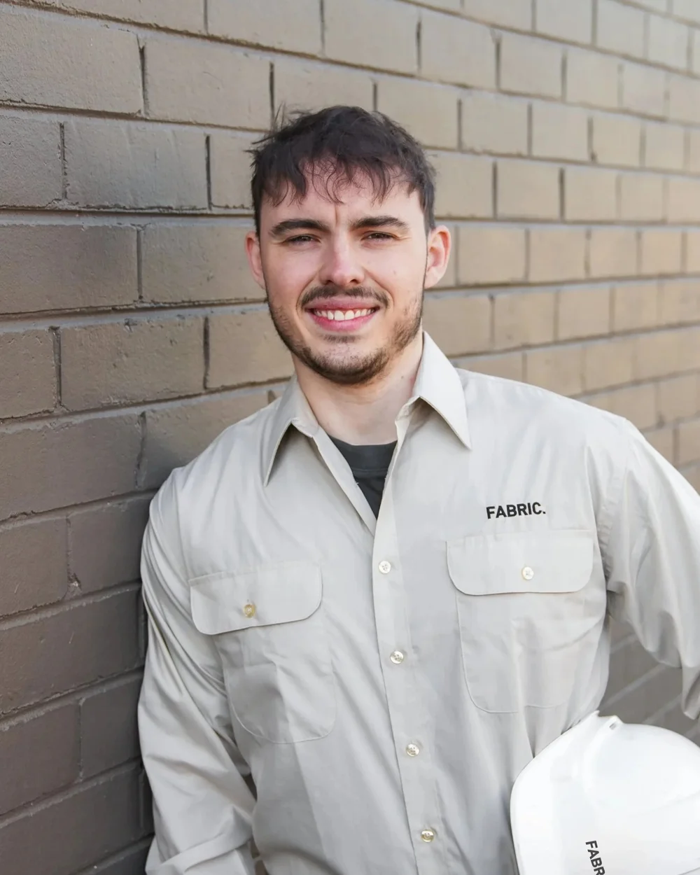 Young man in a beige work shirt holding a hard hat, standing by a brick wall.