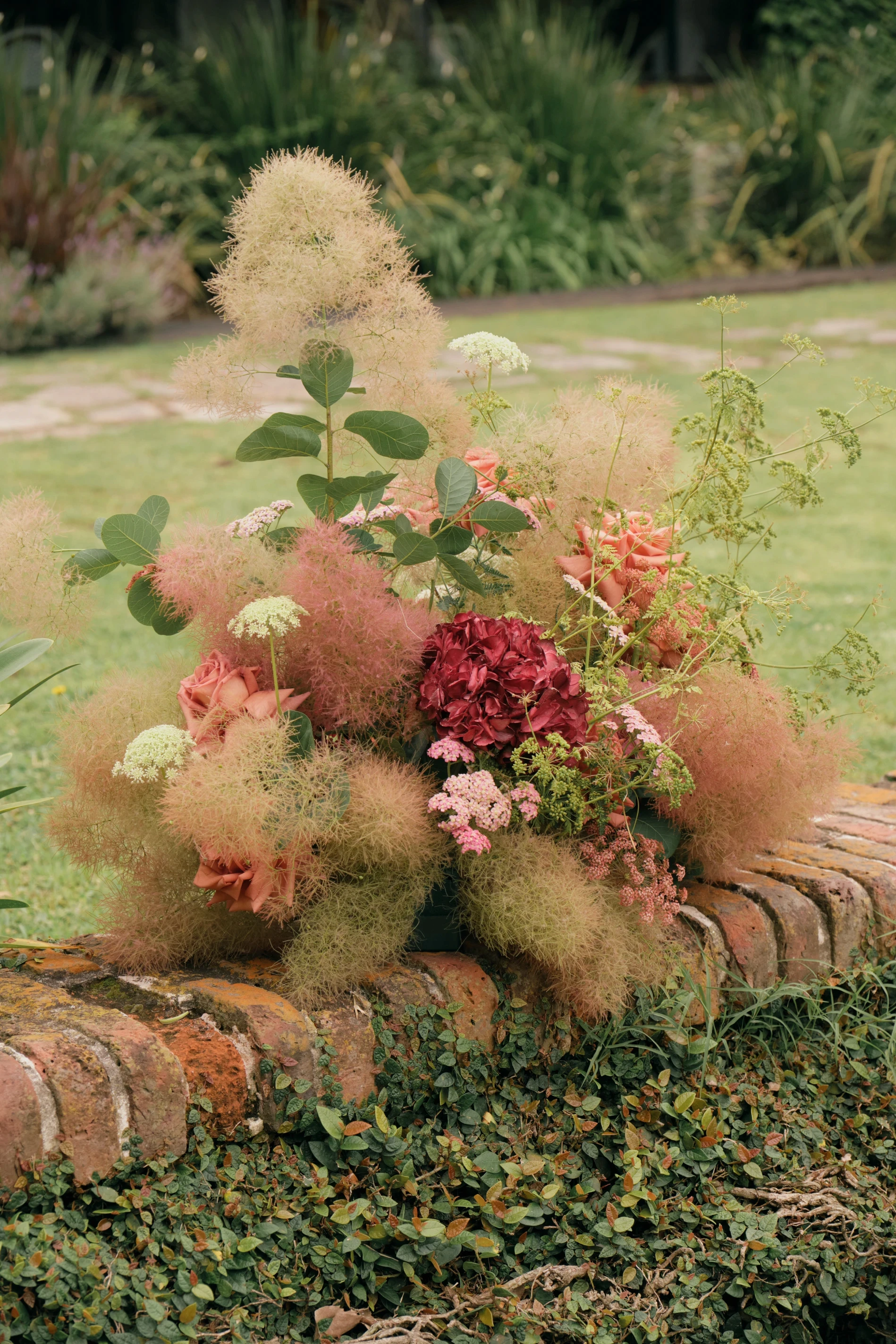 Centro de mesa silvestre con hortensia bordó y smoke bush sobre un muro de ladrillo.
