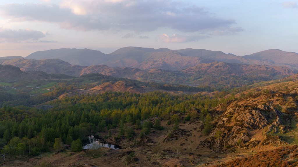 A wide vista of mountain ranges seen from Holme fell. A forest in the foreground with the unused reservoir visible.