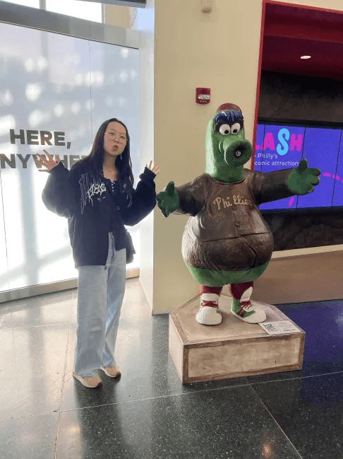 Una persona con una camiseta negra y pantalones de color claro está de pie en la cima rocosa de una montaña al atardecer, sosteniendo una cámara frente a su cara para tomar una foto. Un mar de nubes se extiende detrás de ellos, iluminado por la luz dorada del sol poniente.