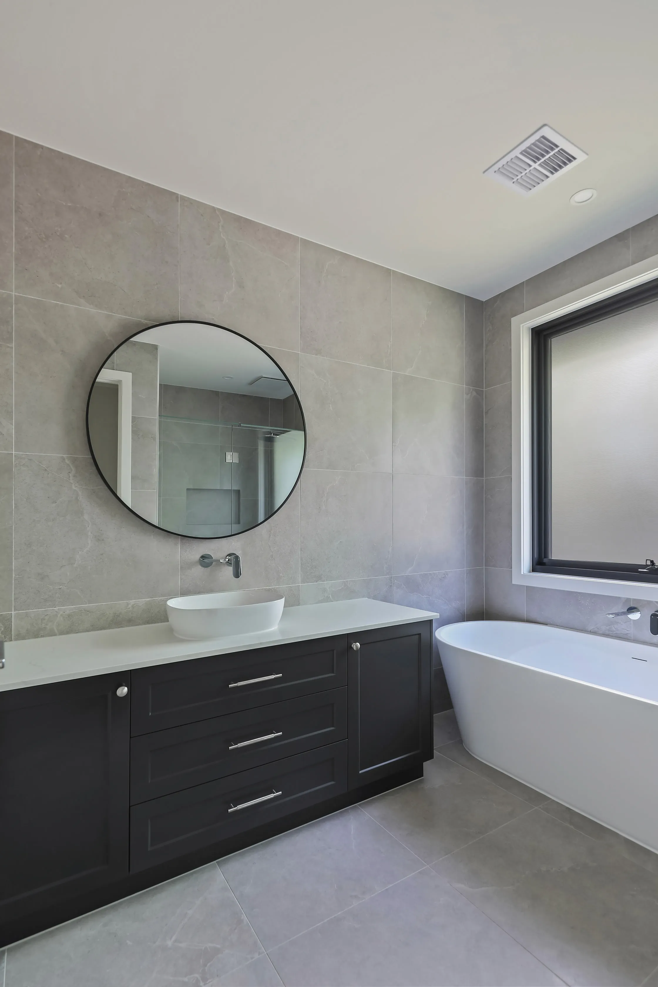 Bathroom in grey stone-look tiles with a round black-framed mirror, white vessel basin on a charcoal shaker vanity with white stone top, a freestanding bath and a frosted black-framed window